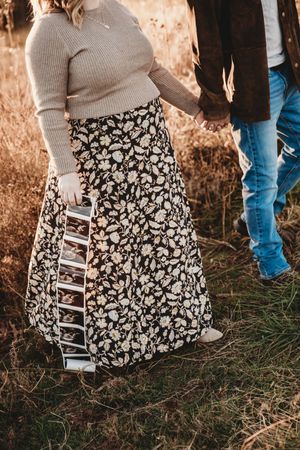 Couple holding hands in autumn field, pregnant woman in floral skirt displaying ultrasound photos at golden hour.