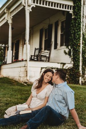 Smiling couple sits on lawn in front of charming white Victorian farmhouse, man whispers to woman holding book.
