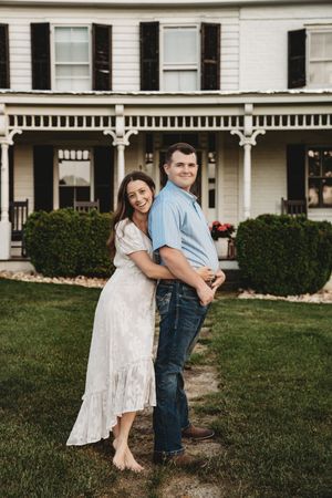 A smiling couple embraces on a green lawn in front of a charming white Victorian farmhouse with black shutters.