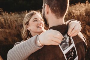 Smiling woman embracing her partner outdoors, holding an ultrasound photo during a golden hour pregnancy reveal.