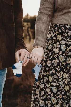 Couple holding tiny baby socks together, pregnancy announcement, wearing floral skirt and brown suede jacket outdoors.