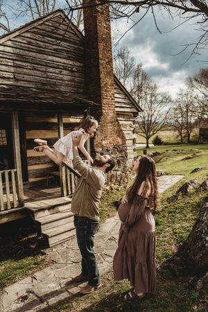 Pregnant woman watches man lift young girl outside rustic log cabin during family maternity photo session.