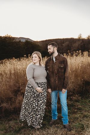 Couple standing in a golden field at sunset, woman in floral skirt and gray sweater, man in brown jacket and jeans.