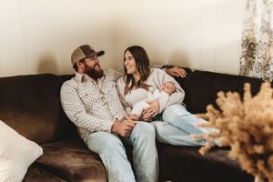 A couple sits on a dark sofa with their newborn baby, smiling warmly in a cozy home setting.