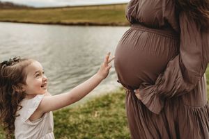 A young girl smiles while touching a pregnant woman's belly near a serene lake outdoors.