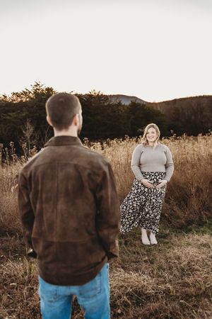 Pregnant woman in floral skirt cradling bump smiles at man in brown jacket in golden meadow at sunset.