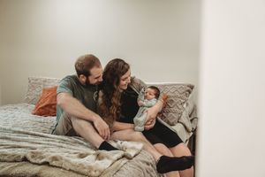 New parents sitting on bed together, lovingly gazing at their newborn baby wrapped in a blanket.