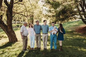 Multi-generational family of six poses together outdoors under trees on lush green grass during a sunny day.