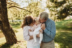 Grandparents kissing a baby girl outdoors in a sunny green park, surrounded by trees in warm natural light.
