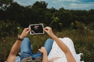 Couple lying in a field holding an ultrasound photo, pregnancy announcement outdoors.