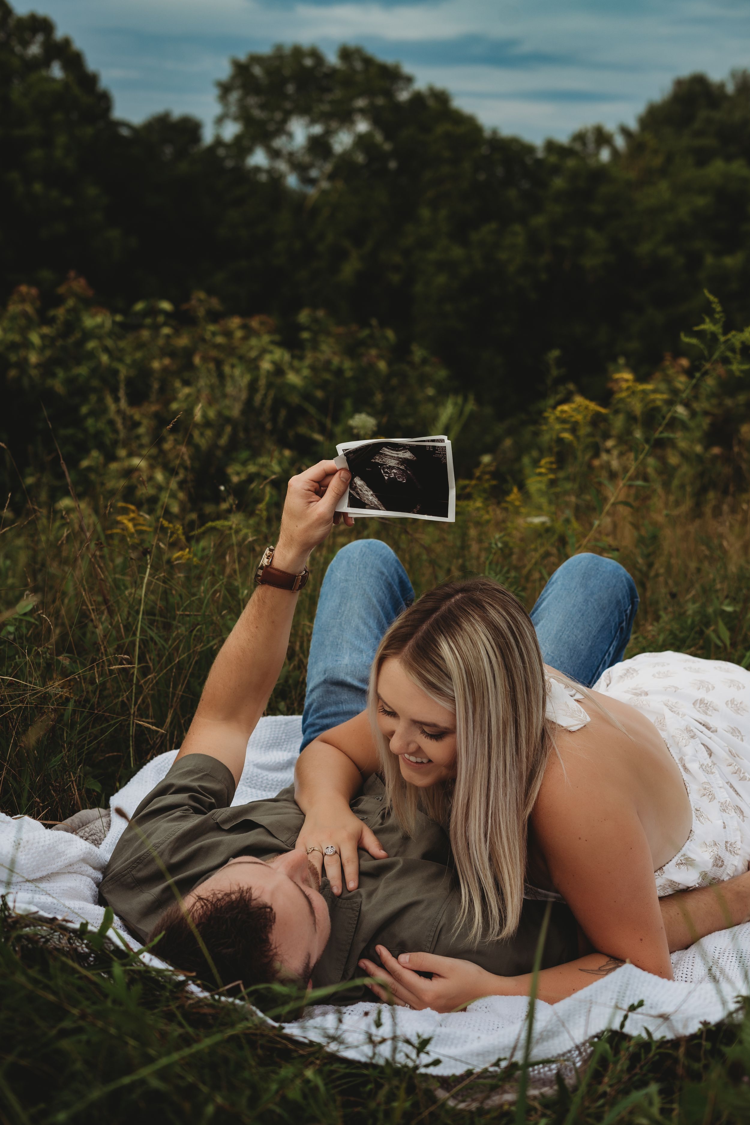 Couple lying on a blanket in a meadow, smiling and holding an ultrasound scan photo.