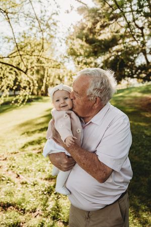 Elderly grandfather kissing smiling baby girl outdoors in sunny green park setting.