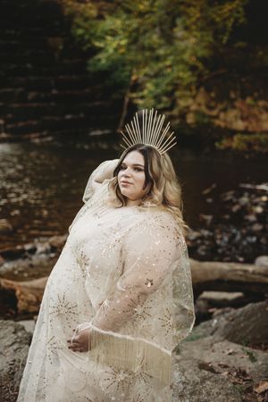 Plus-size bride in beaded lace gown and gold sunburst crown poses by rocky creek in autumn forest.
