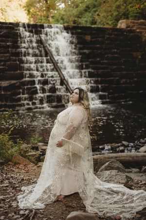 Plus-size bride in lace gown and crown poses gracefully before a serene waterfall in a natural outdoor setting.