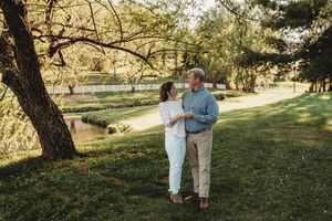 Couple holding hands and dancing together in a lush green park near a pond on a sunny day.