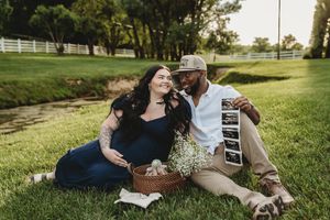 Pregnant couple sitting on grass at a farm, holding ultrasound photos and a flower basket during a maternity photoshoot.