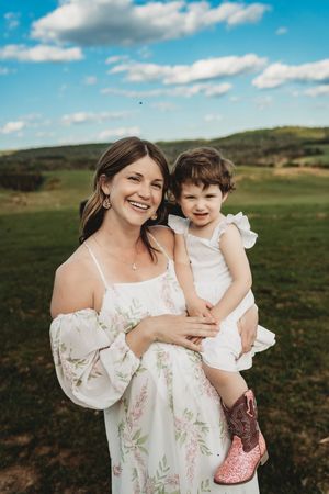 Smiling woman in floral dress holds young girl in white dress and pink cowboy boots outdoors.