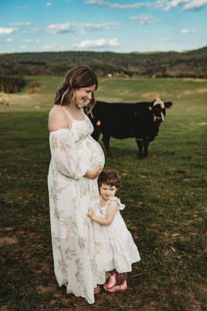 Pregnant woman in floral dress with toddler girl in white dress standing in green field with black cow in background.