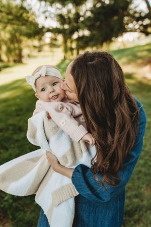 A mother tenderly kisses her smiling baby girl outdoors, both dressed in soft pastel tones against a lush green background.