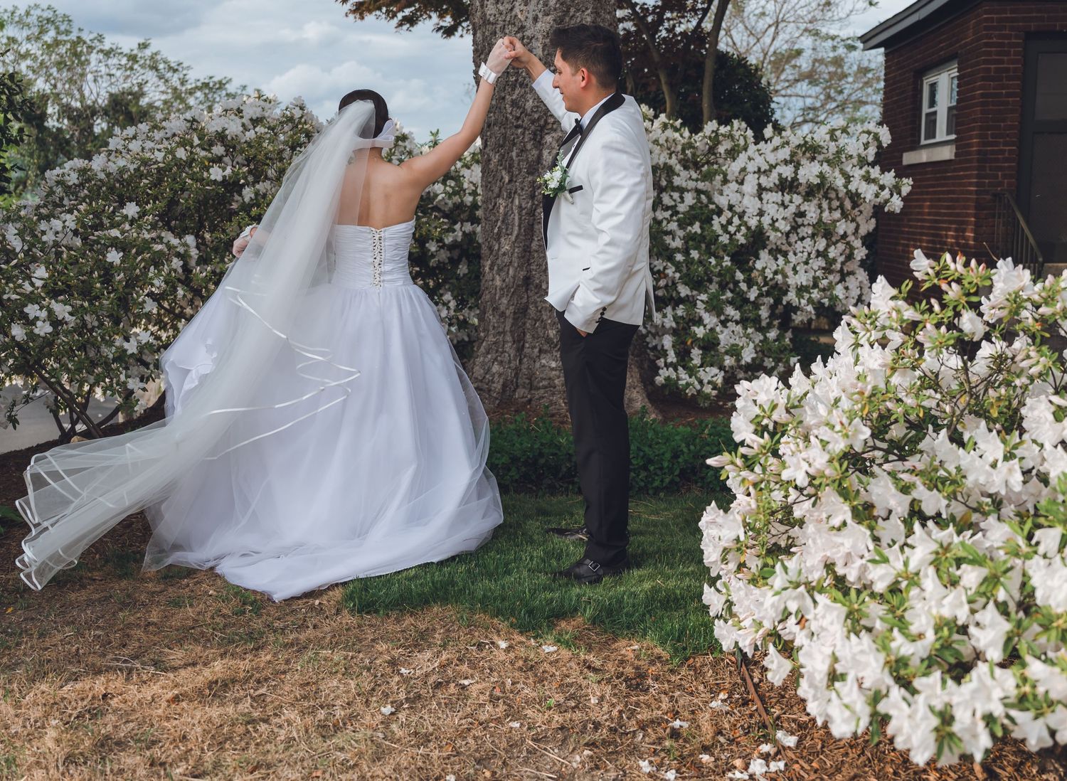 Bride and groom dancing outdoors near blooming white flowers and trees on their wedding day.
