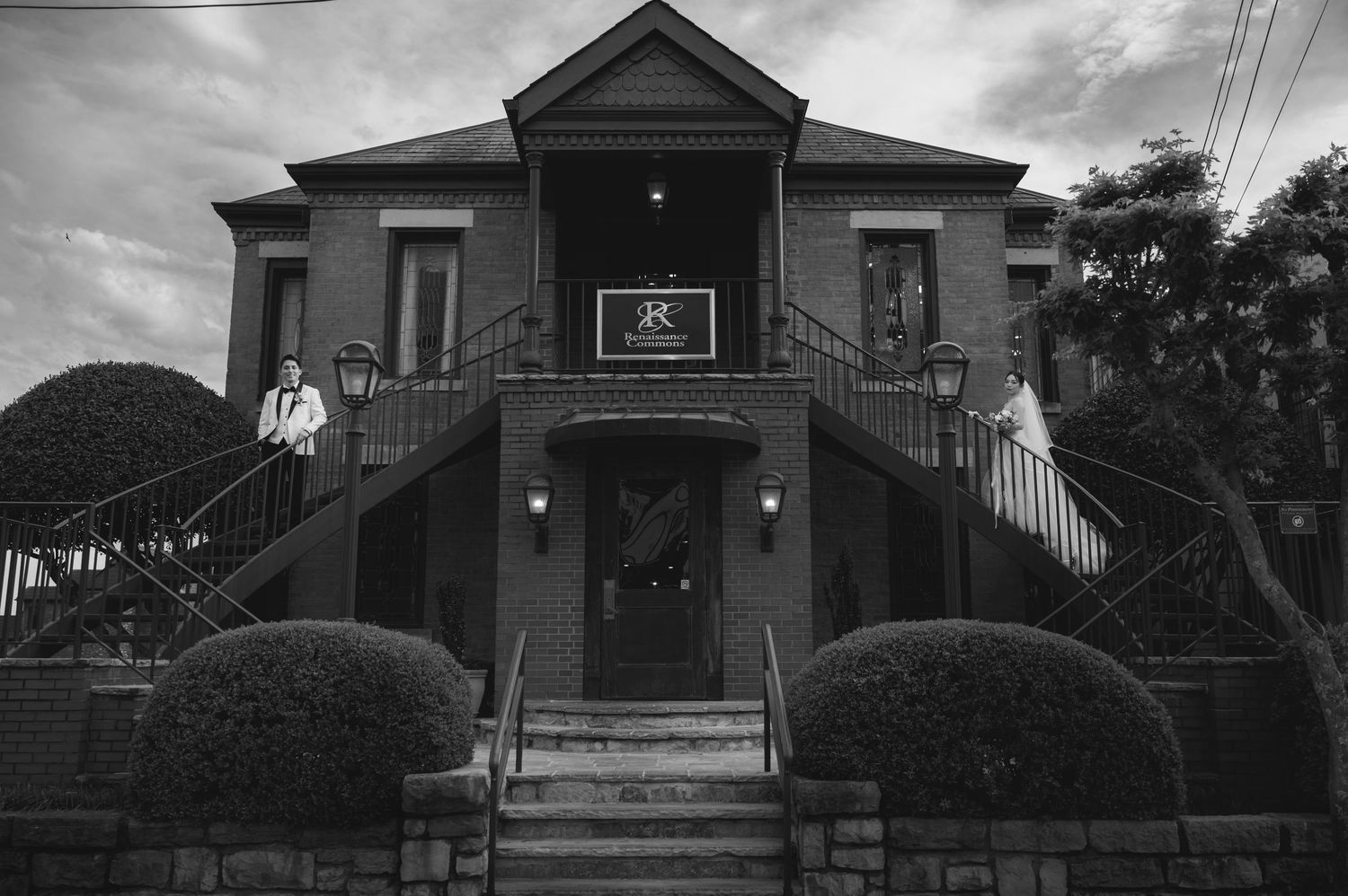 Black and white photo of an elegant two-story brick building with dual staircases and manicured hedges.