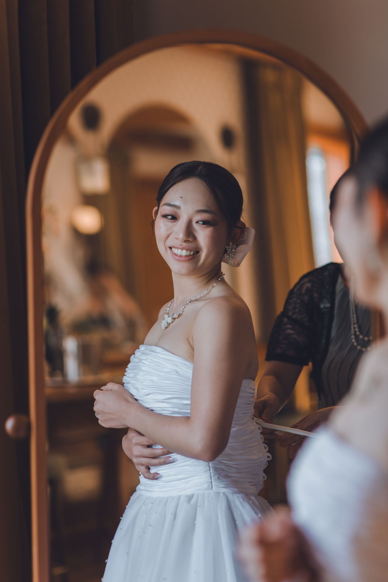 Smiling Asian bride in strapless white wedding dress standing before arched mirror while being assisted by attendant.