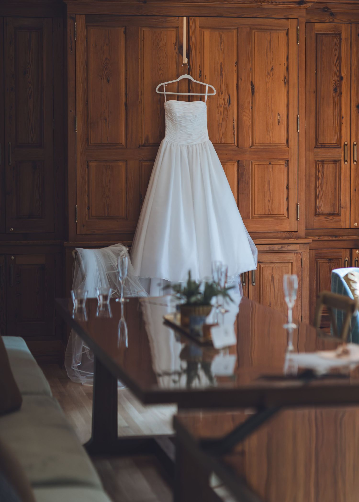 White wedding dress hanging on wooden wardrobe in elegant room with dining table and champagne glasses.
