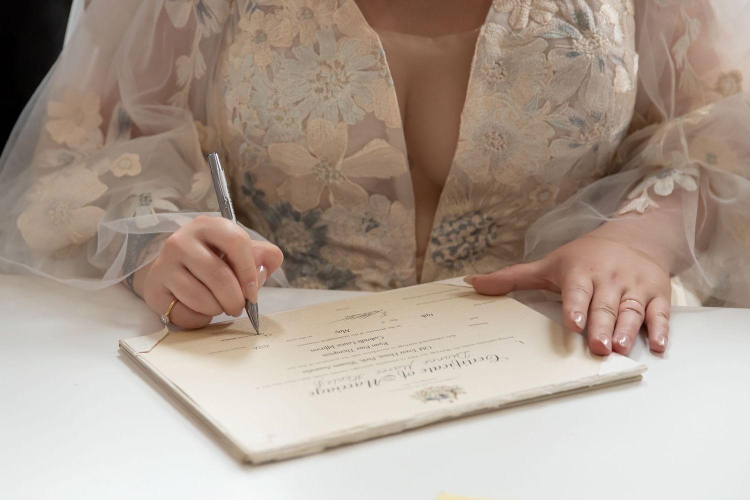 Bride in floral gown signing wedding certificate at white table, wearing ring on finger.