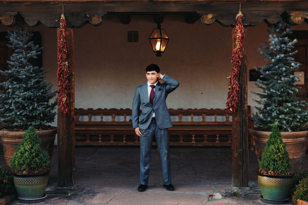 Groom in gray suit stands between red chili pepper ristras and evergreen trees on a rustic adobe porch.