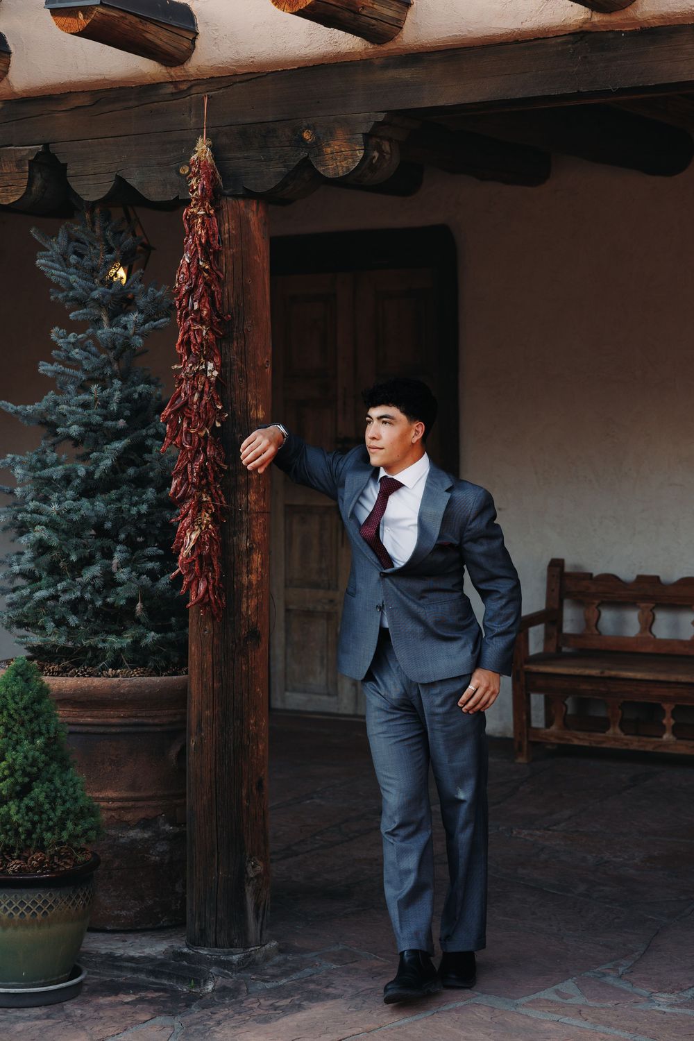 Young man in gray suit and tie posing by rustic wooden pillar with hanging dried chili peppers on a Southwest-style porch.