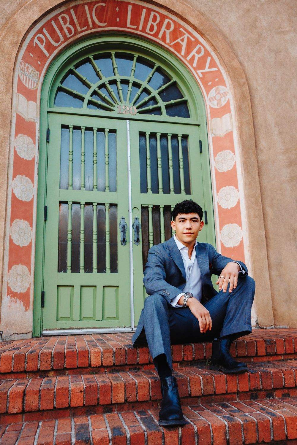 Young man in blue suit sits on brick steps before a green arched Public Library entrance door.
