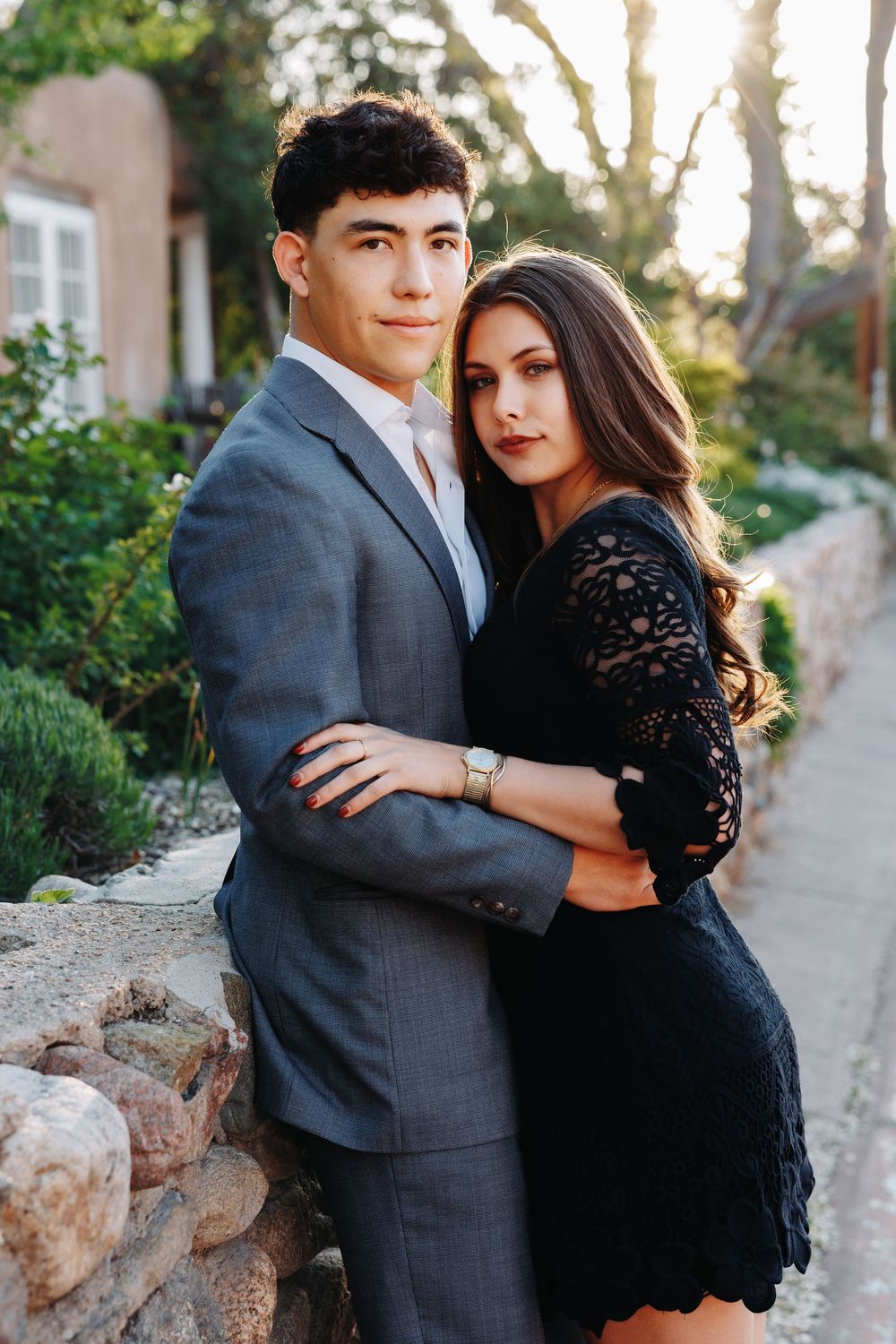A young couple in formal attire poses outdoors, the man in a gray suit and woman in a black lace dress, embracing near a stone wall.
