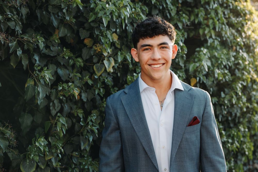Young man in gray blazer and white shirt smiling outdoors against lush green ivy wall.