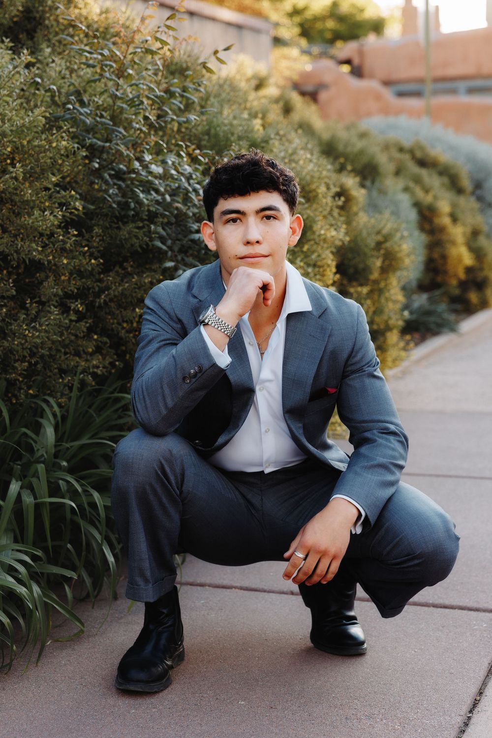 Young man in gray suit crouching thoughtfully on pathway with hand on chin near green hedges.