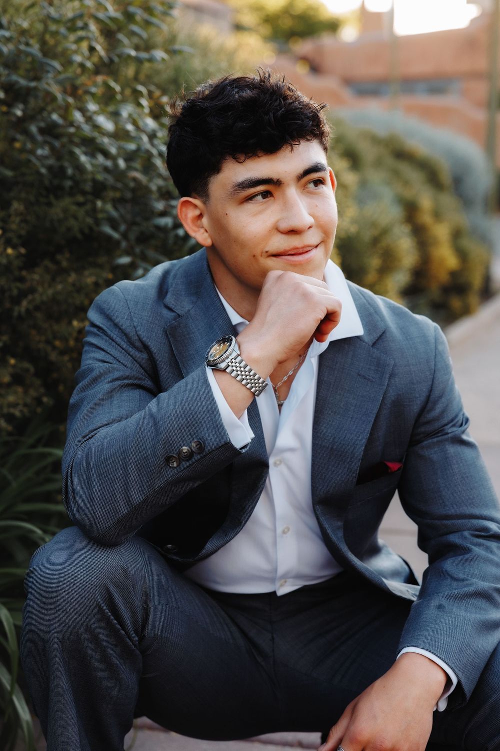 Stylish young man in gray suit with watch sitting outdoors, resting chin on hand in thoughtful pose near green shrubs.