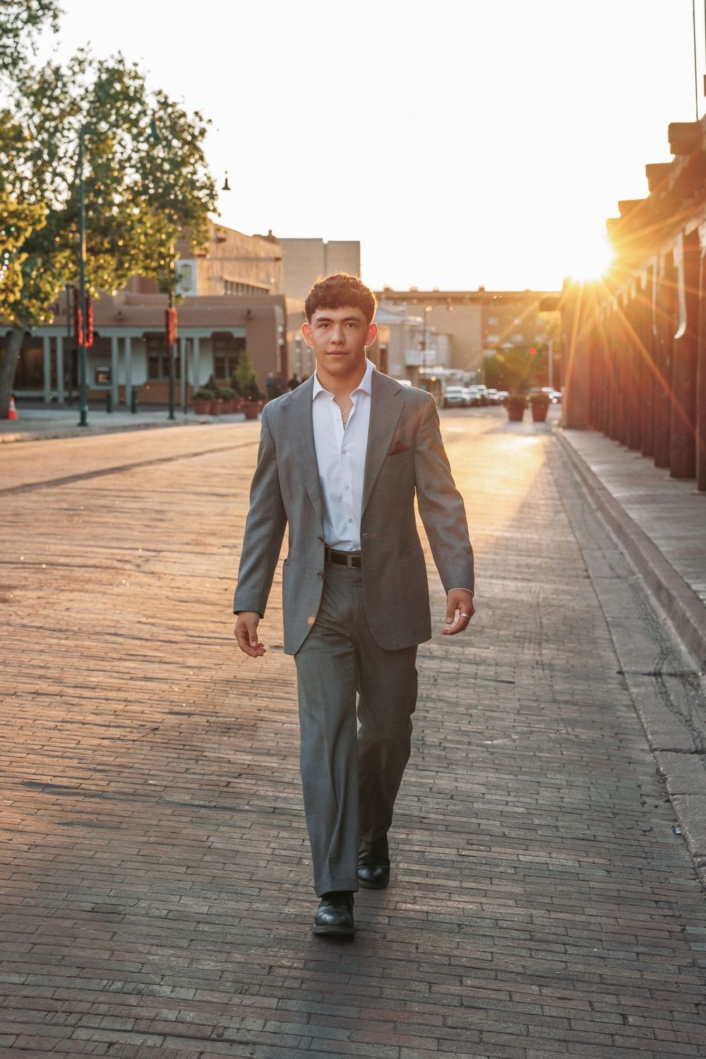 Young man in gray suit walking toward camera on brick street at golden hour sunset.