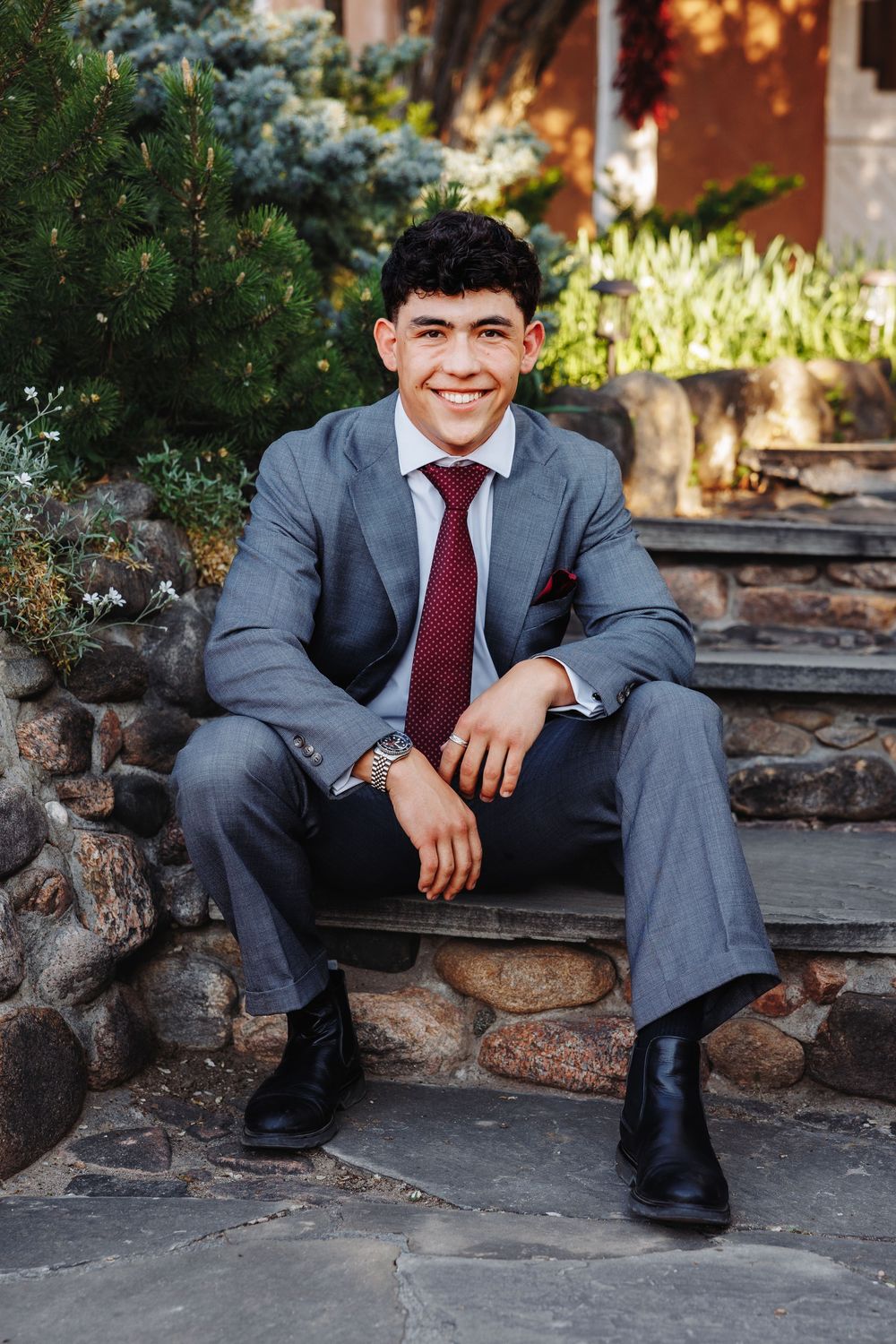 Young man in gray suit and burgundy tie sitting on stone steps, smiling confidently outdoors.