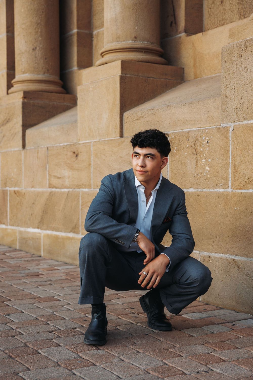 Young man in gray suit crouching by stone building columns on brick pavement, looking confidently at camera.