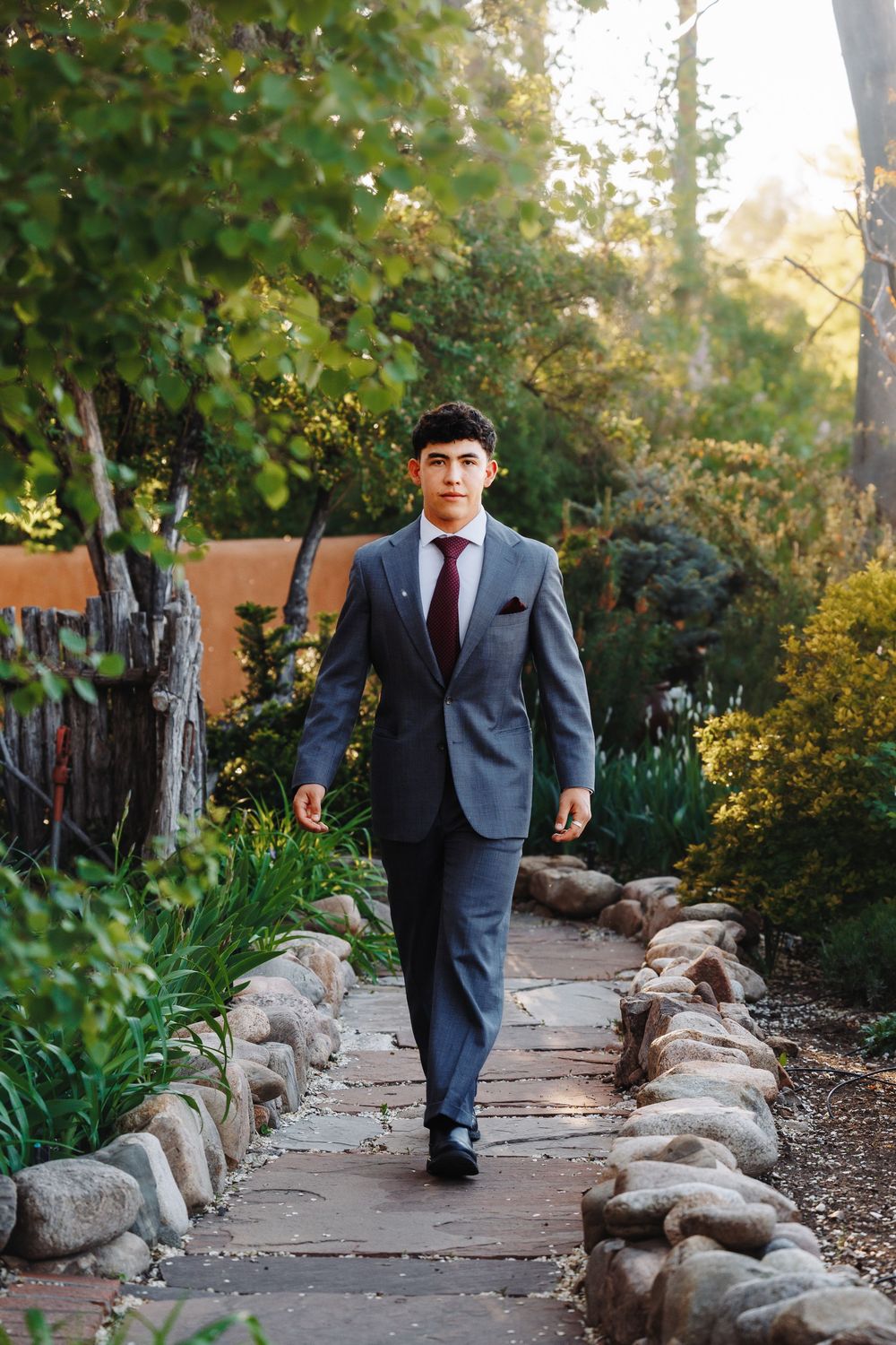 Young man in gray suit and burgundy tie walks along a stone garden path surrounded by lush greenery.