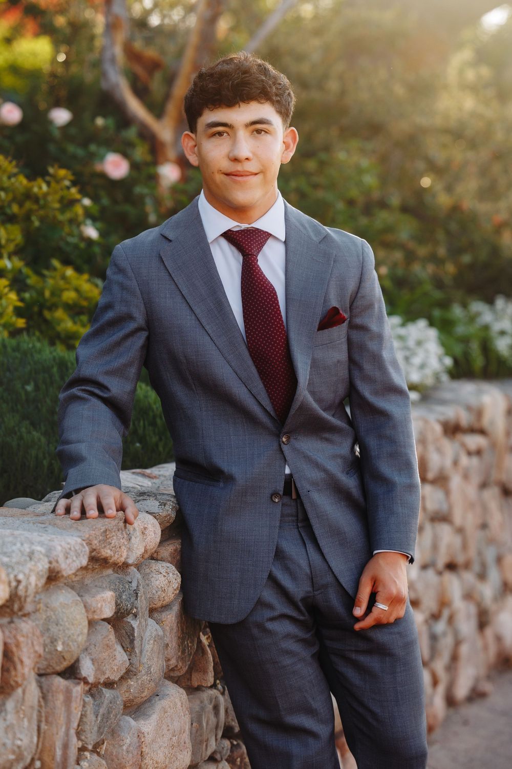 Young man in gray suit with burgundy tie and pocket square posing outdoors near stone wall with flowers.