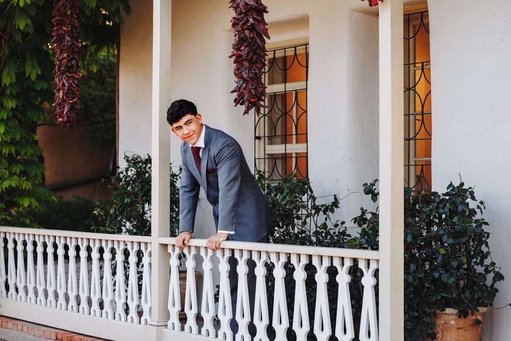 Groom in gray suit leans on white porch railing outside decorated building with red chili peppers.