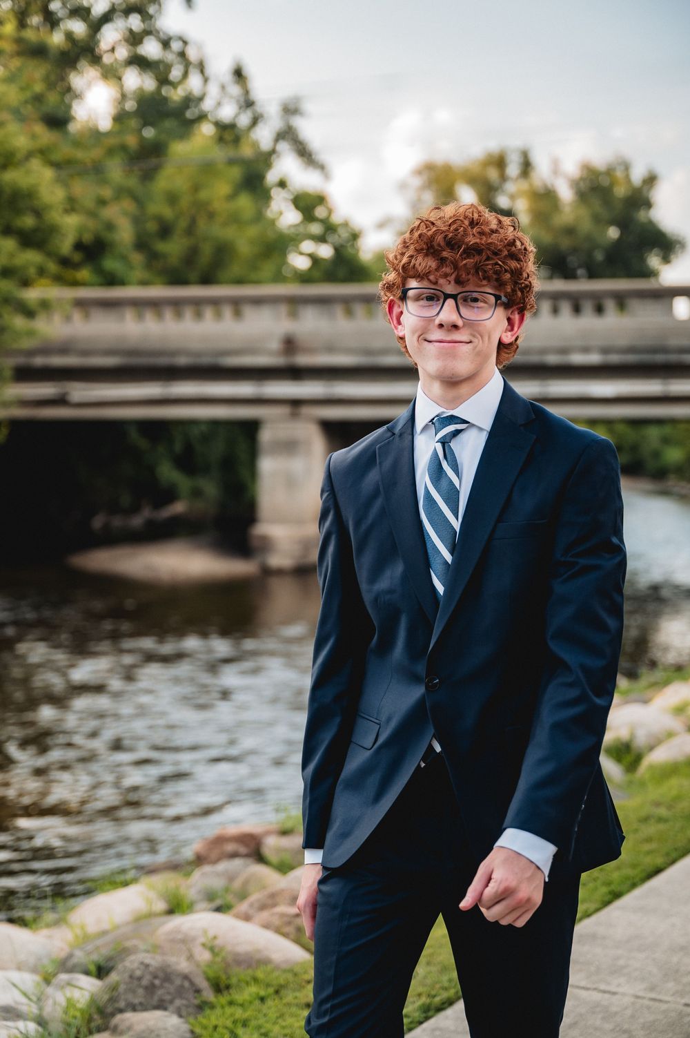 Young man with curly red hair wearing a navy suit and striped tie, smiling outdoors near a river and bridge.
