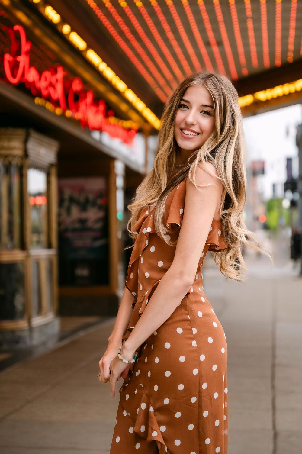 Young woman in brown polka dot dress smiling on city sidewalk near vintage theater marquee lights.