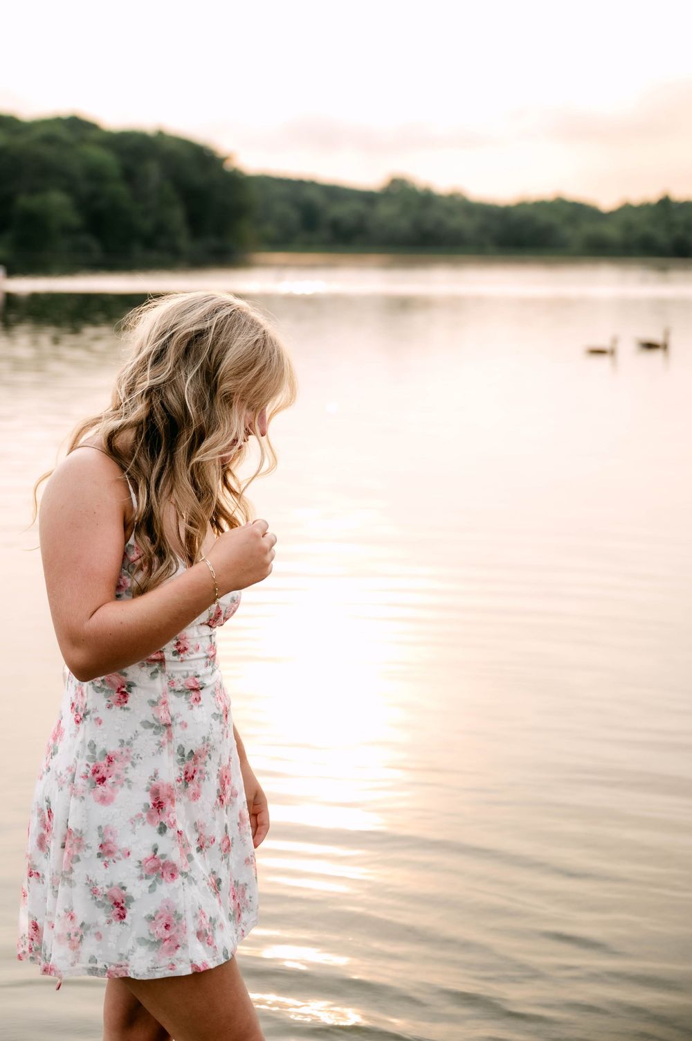 Young blonde woman in floral dress standing by calm lake at golden sunset.
