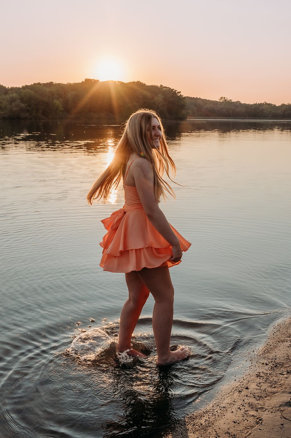 Young blonde woman in peach dress wading in river at golden sunset, hair flowing, smiling over shoulder.