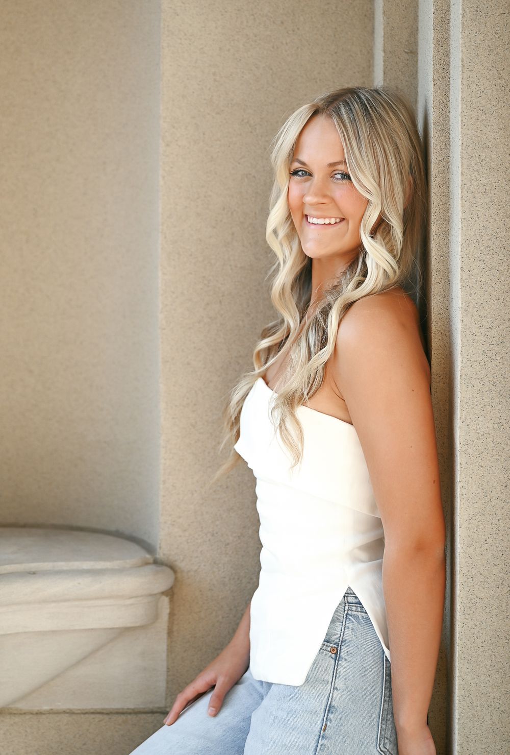 Smiling blonde woman in white strapless top and jeans leaning against a stone pillar outdoors.