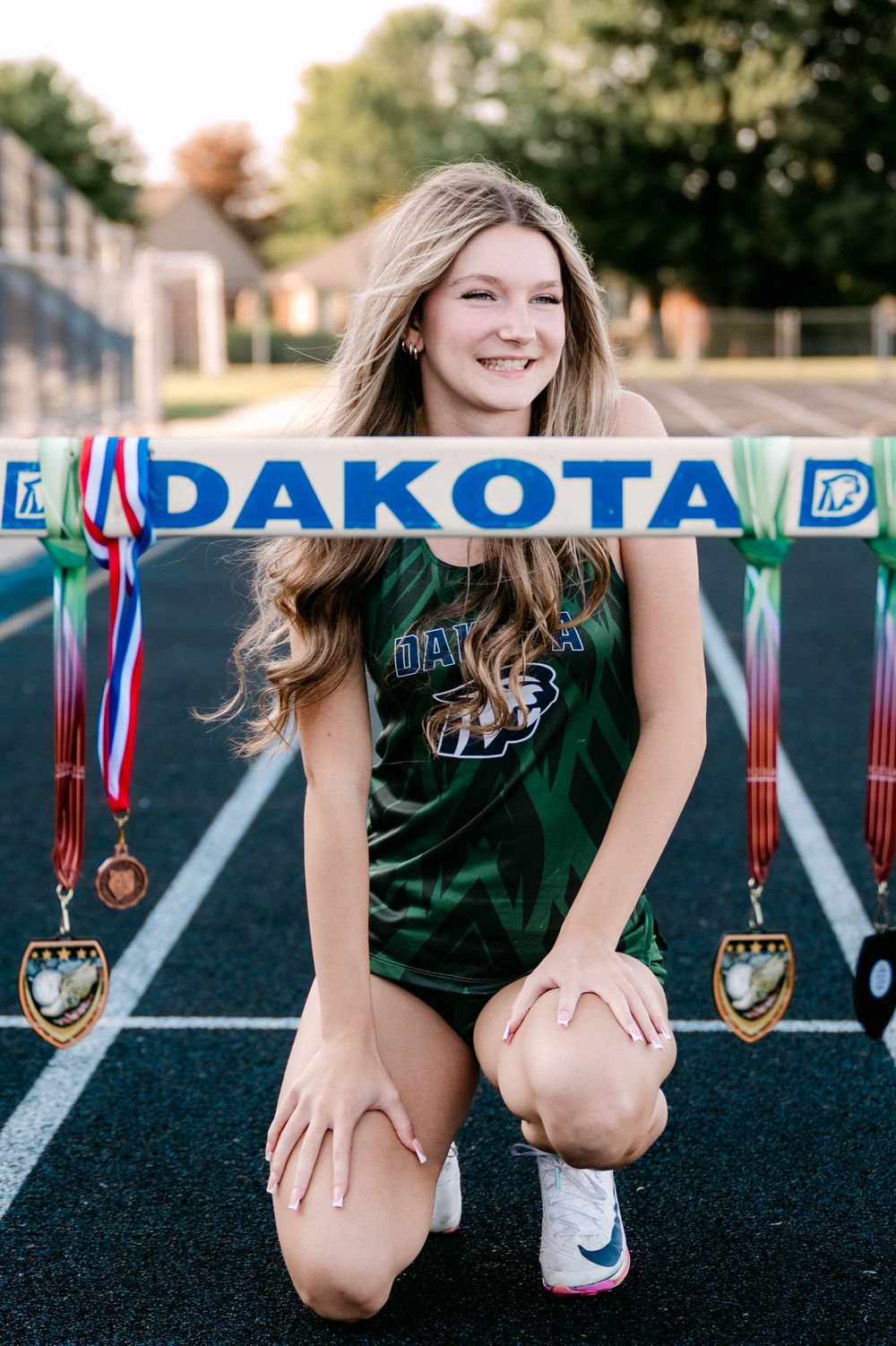 Smiling teen athlete kneeling on track with Dakota banner and medals for senior photos.