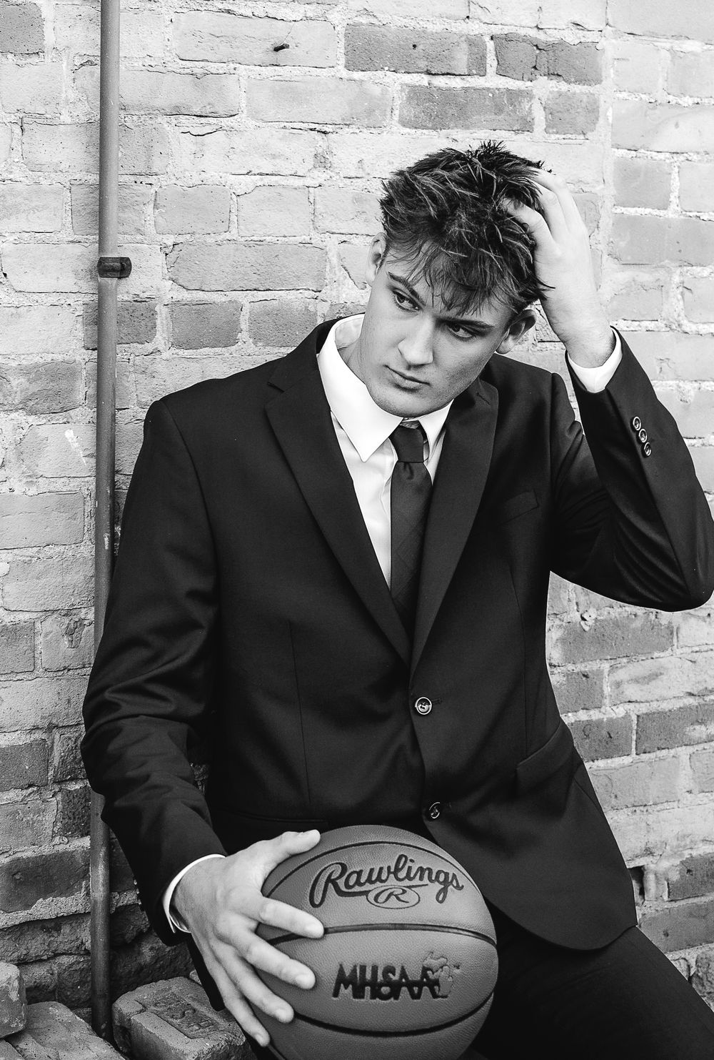 Young man in black suit and tie holding a Rawlings basketball, posing against a brick wall in a black and white photo.