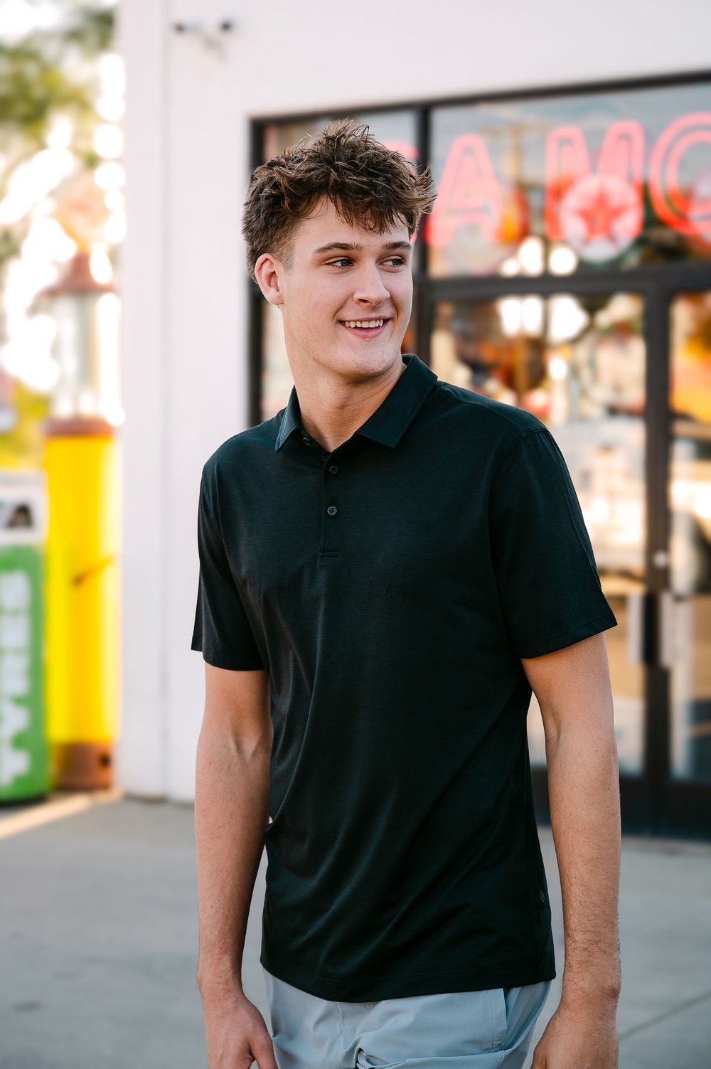 Young man smiling in a black polo shirt standing outside a store with neon signs.