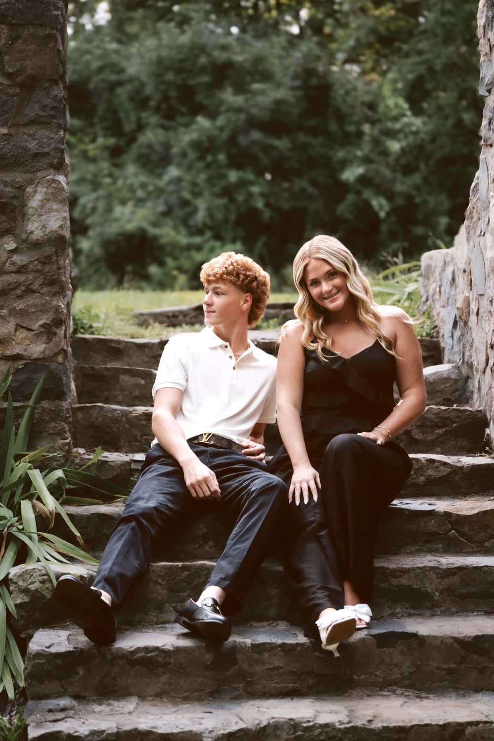 Two teens sit on stone steps outdoors, boy in white polo and girl in black outfit, smiling together near lush greenery.